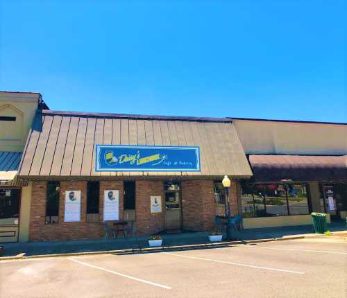 Exterior of a café named "Daisy's Lemonade," featuring a blue sign and a sunny sky. Parking lot in front.