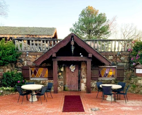 A rustic stone building with a wooden door, surrounded by greenery and two outdoor tables on a brick patio.