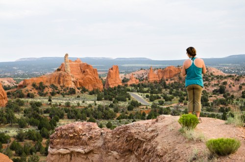 A person stands on a rocky ledge, overlooking a vast landscape of red rock formations and greenery under a cloudy sky.