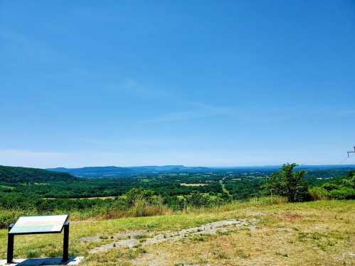 A panoramic view of green valleys and distant mountains under a clear blue sky, with a sign in the foreground.