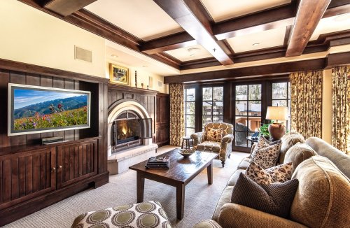 Cozy living room with a fireplace, wooden beams, a TV, and comfortable seating, featuring large windows and decorative curtains.