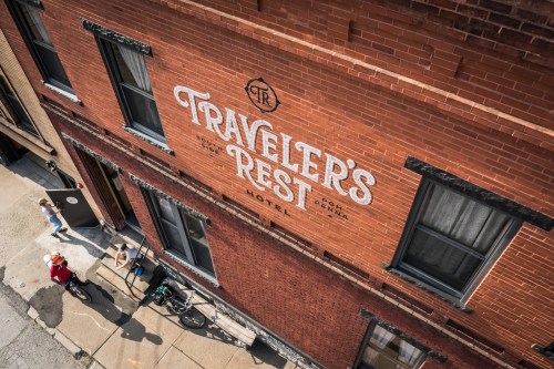 Aerial view of a brick building with "Traveler's Rest Hotel" painted on the wall, featuring a bicycle and people outside.