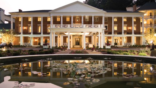 A grand brick building with white columns, reflecting in a pond with lily pads, illuminated at dusk.