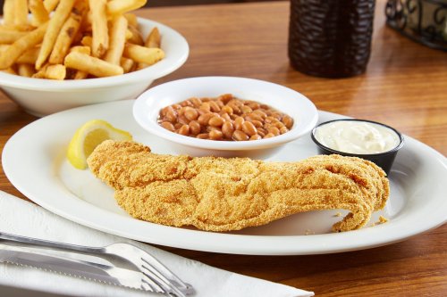 A plate with a fried fish fillet, lemon slice, baked beans, and a side of dipping sauce, with fries in the background.