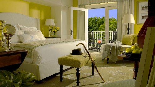 Brightly lit bedroom with yellow walls, a large bed, and a balcony view. Decor includes a cane and green apples.