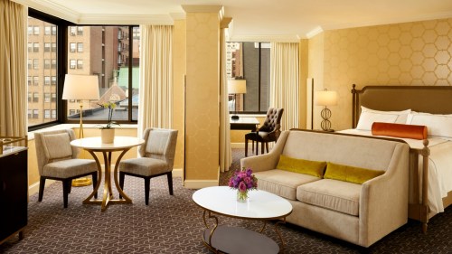 A stylish hotel room featuring a bed, seating area, table, and large windows with city views.