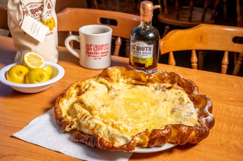 A large, fluffy pancake on a plate, with a cup of coffee, a bottle of syrup, and a bowl of lemons in the background.