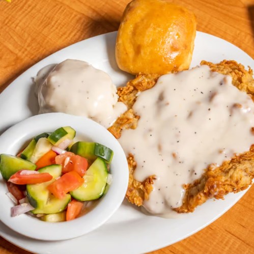 A plate featuring fried chicken with gravy, a biscuit, mashed potatoes, and a side salad of cucumbers and tomatoes.