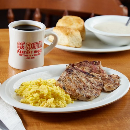 A plate with grilled pork chops, scrambled eggs, a cup of coffee, and biscuits with gravy in the background.