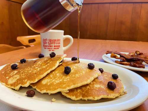 A plate of pancakes topped with blueberries, with syrup being poured, alongside a cup of coffee and a plate of bacon.