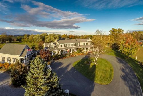 Aerial view of a charming country inn surrounded by trees and a circular driveway under a blue sky with clouds.