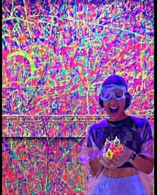 A person in protective gear holds colorful paint bottles in front of a vibrant, abstract painted wall.