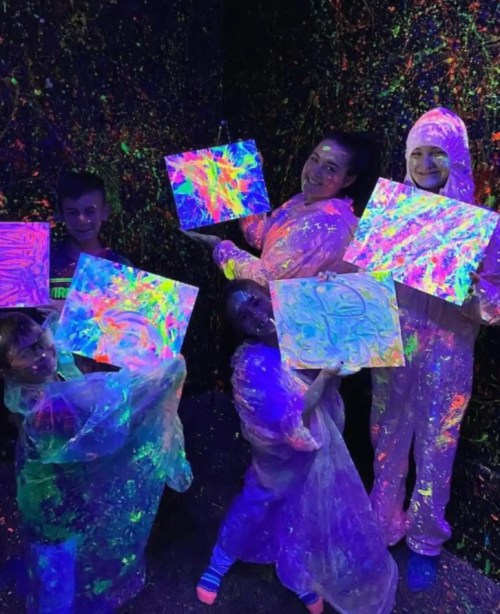 A group of five people in glowing outfits hold colorful, neon-painted canvases in a dark, UV-lit room.