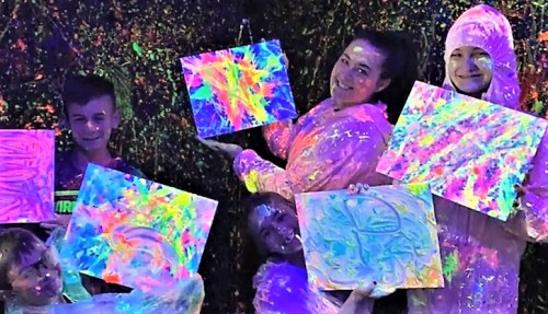 A group of five people in colorful outfits hold vibrant, neon paintings against a splattered background.