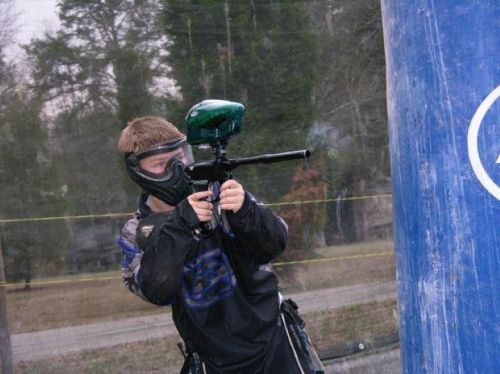 A person in a mask aims a paintball gun while crouching behind a blue barrier in an outdoor setting.