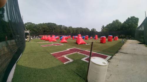 A paintball field with red and blue inflatable obstacles on green turf, surrounded by trees and a black netting.