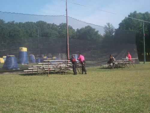 Two players in pink gear prepare for a paintball game near a netted field with colorful obstacles and spectators on benches.