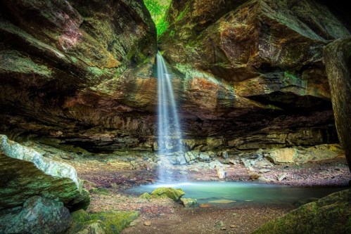 A serene waterfall cascades from a rocky overhang into a tranquil pool surrounded by lush greenery.