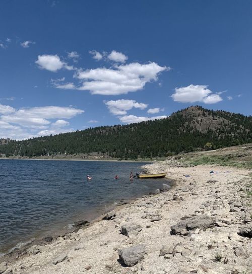 A serene lakeside scene with a yellow boat, people wading in the water, and mountains in the background under a blue sky.