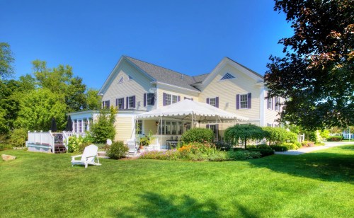 A charming yellow house with a large tent, surrounded by lush greenery and colorful flowers on a sunny day.