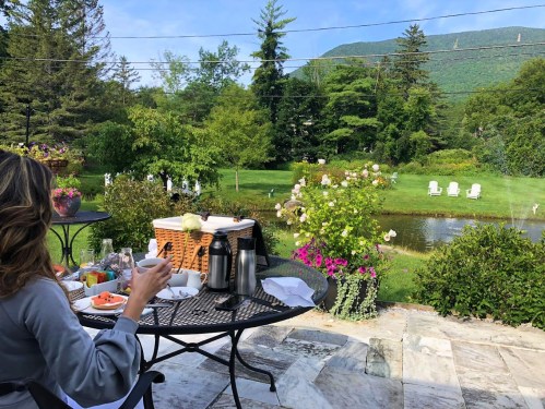 A person enjoys breakfast on a patio, overlooking a lush garden and pond with mountains in the background.