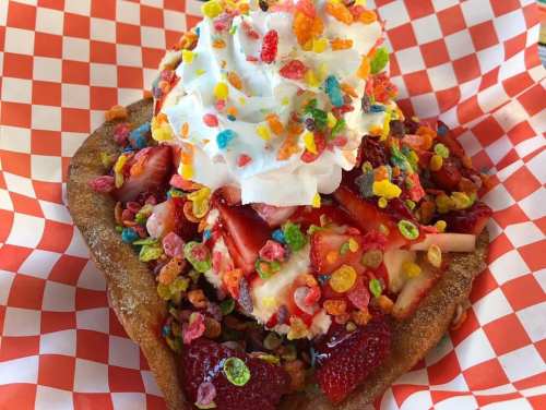 A dessert featuring a fried dough base topped with ice cream, strawberries, whipped cream, and colorful cereal.