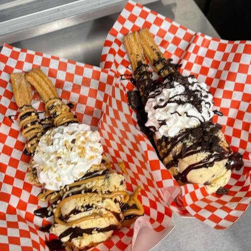 Two servings of churros topped with ice cream, whipped cream, chocolate sauce, and crushed cookies in checkered baskets.