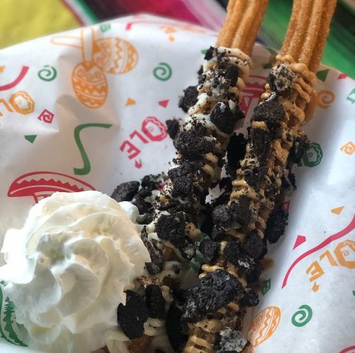 Two churros topped with crushed cookies and drizzled with cream, served with a dollop of whipped cream on a colorful paper.