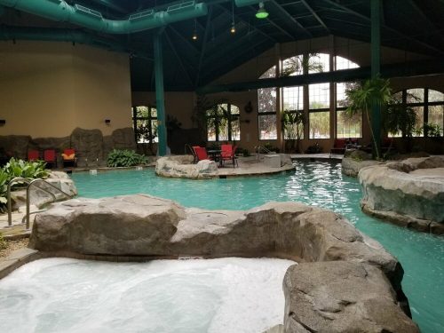 Indoor pool area with rocky features, hot tubs, and tropical plants, surrounded by large windows and lounge chairs.