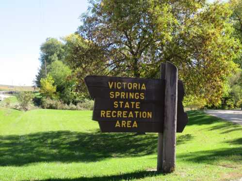 Wooden sign for Victoria Springs State Recreation Area, surrounded by green grass and trees on a sunny day.