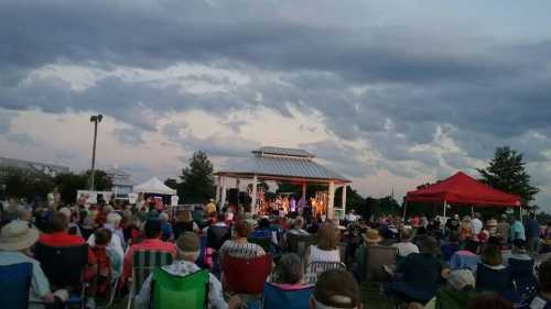 A large crowd gathers in an outdoor setting for a performance, with a stage and colorful tents under a cloudy sky.