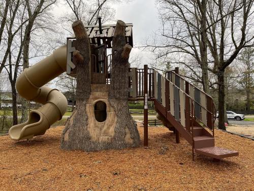 A playground structure resembling a tree with a slide and stairs, surrounded by mulch and trees in a park setting.