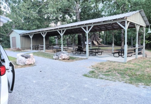 A covered picnic area with multiple tables, surrounded by trees and gravel pathways.