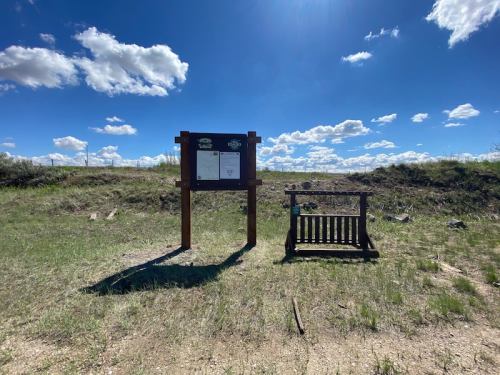 A wooden signboard stands in a grassy area under a blue sky with clouds, next to a small bench.