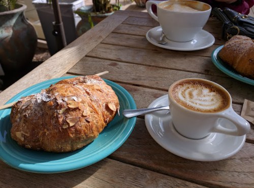 A croissant topped with almonds next to a cup of latte on a wooden table.
