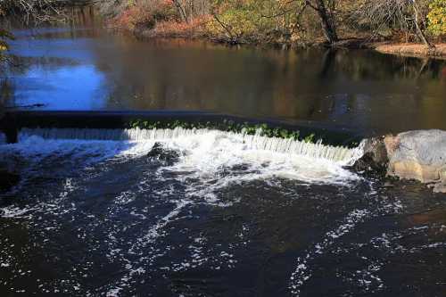 A serene river scene with a small waterfall cascading over a dam, surrounded by autumn foliage.