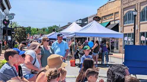 A crowd watches an outdoor event with tents and shops in a lively street scene on a sunny day.