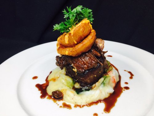 A gourmet dish featuring tender beef, creamy mashed potatoes, onion rings, and a garnish of parsley on a white plate.
