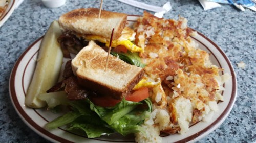 A plate with a sandwich, lettuce, tomato, pickle, and crispy hash browns on the side.