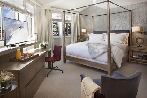 A cozy hotel room featuring a four-poster bed, TV, and a view of snowy mountains through large windows.