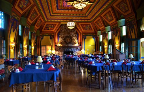 Colorful dining room with blue tables, red napkins, and a stone fireplace, featuring vibrant patterned ceilings and large windows.