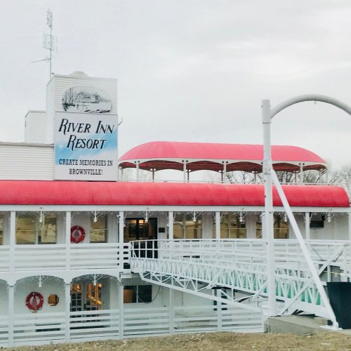 Sign for River Inn Resort in Brownville, featuring a white building with a red awning and a bridge-like walkway.