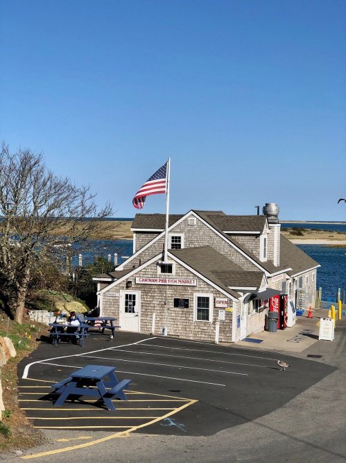 A coastal fish market with an American flag, picnic tables, and a view of the water on a clear day.