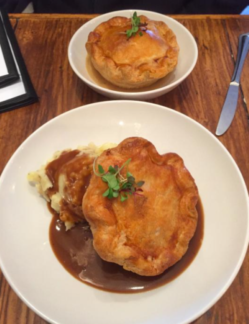 A hearty meat pie served with mashed potatoes and gravy, garnished with a sprig of herbs, on a wooden table.