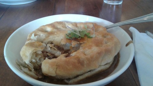 A bowl of savory pie with a flaky crust, filled with meat and topped with fresh herbs, on a wooden table.