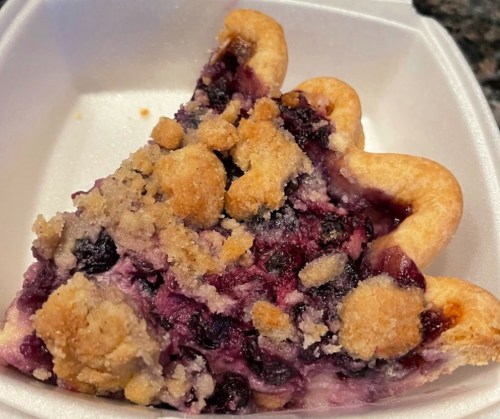 A slice of blueberry pie with a crumbly topping, served in a white takeout container.
