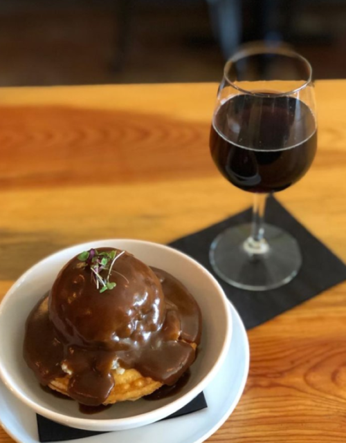 A dessert bowl with a scoop of ice cream topped with chocolate sauce, alongside a glass of dark beverage on a wooden table.