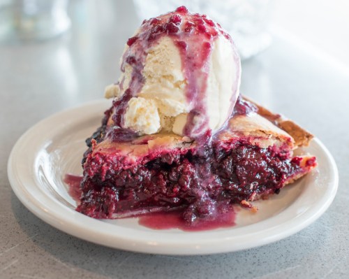 A slice of berry pie topped with melting vanilla ice cream on a white plate.