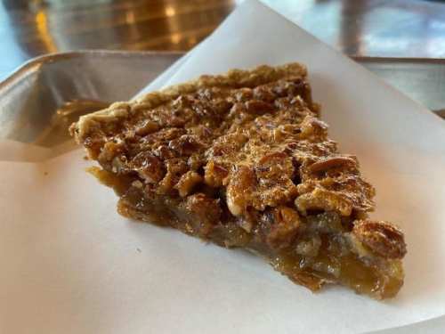 A slice of pecan pie on a white paper, resting on a metal tray with a wooden background.