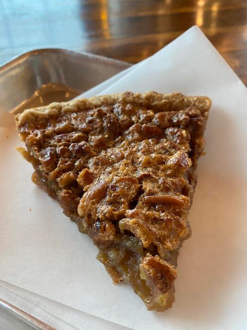 A slice of pecan pie on a white paper, showcasing a golden-brown crust and a rich, nutty filling.